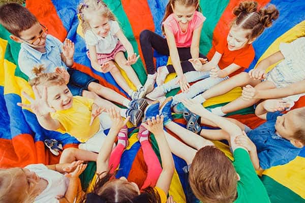 Eine Gruppe Kinder sitzt im Kreis auf einem bunten Fallschirm auf einem Indoor-Spielplatz in Augsburg.